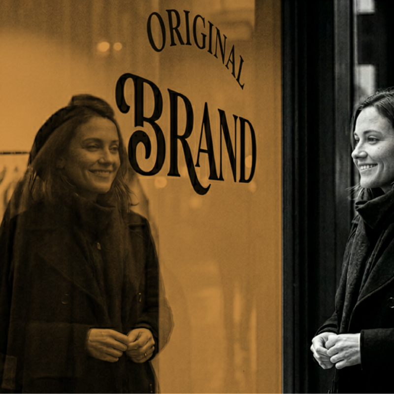 Woman standing outside a shop window with the word “Brand” displayed, representing employer branding, company culture, and attracting the right talent.